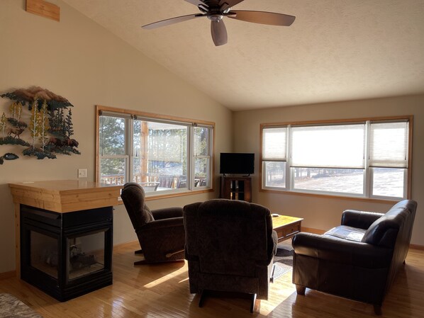 A living room with a fireplace, two armchairs, a sofa, a TV, and large windows offering a view of the outdoors.