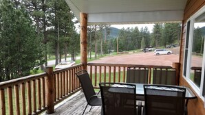 Una terraza de madera con una mesa y sillas, con vista a una carretera con coches aparcados y un área boscosa.
