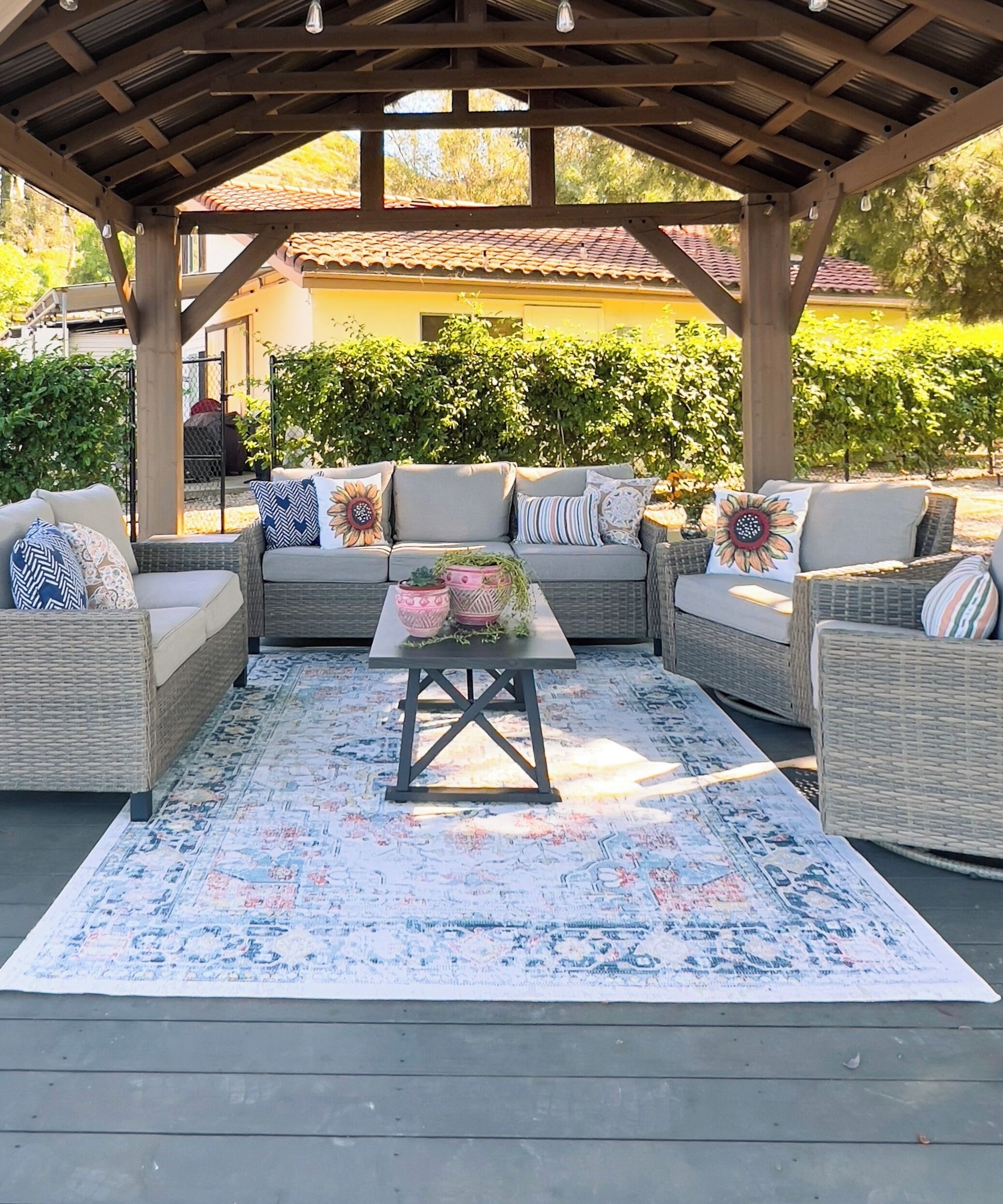 Un espacio de estar al aire libre con mobiliario de mimbre, un tapete estampado y una pérgola.