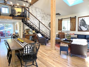 A spacious living area with a wooden staircase, a dining table with chairs, a sofa, and a chessboard.