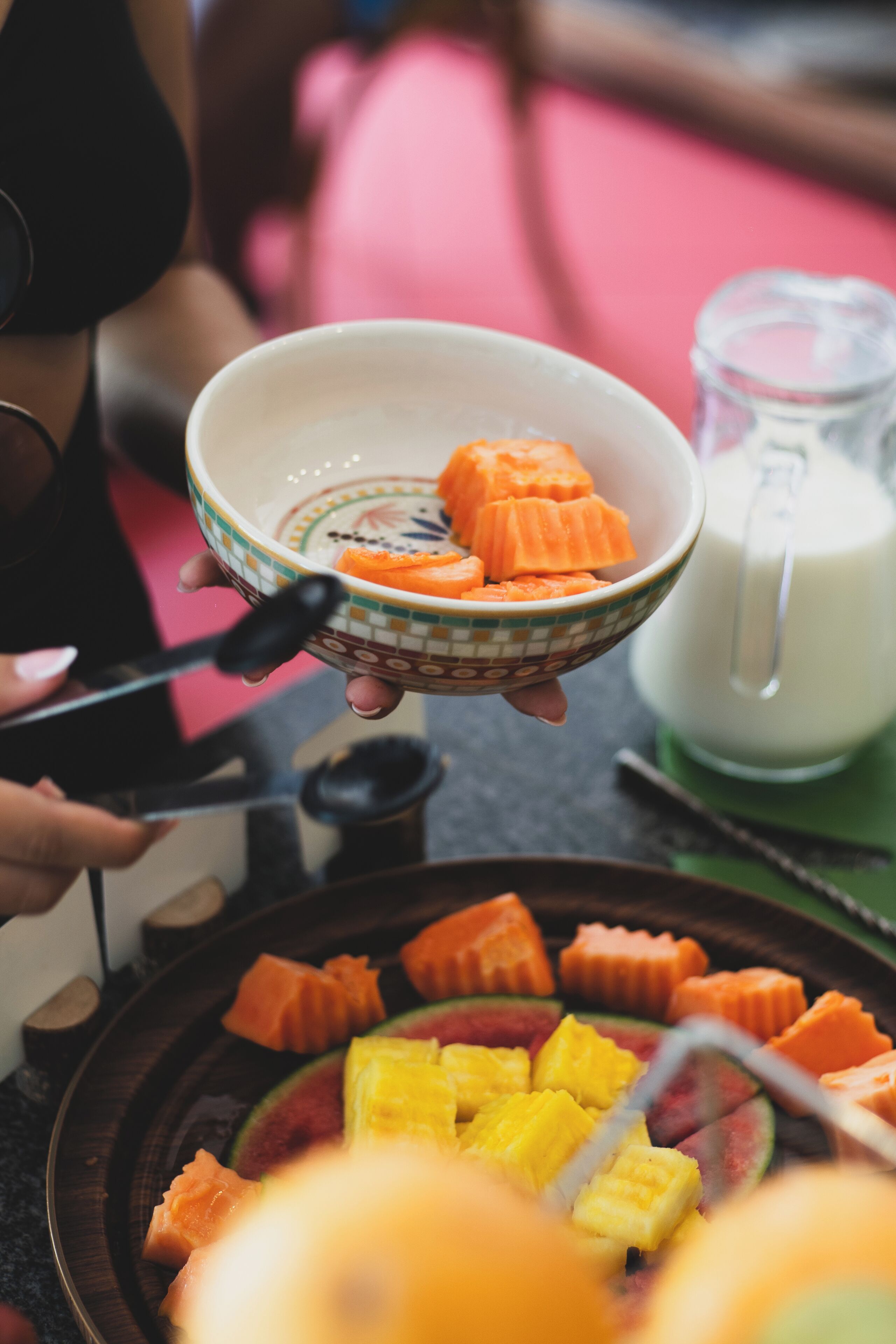 A person holding a bowl with sliced carrots and a glass pitcher of milk on a table with various fruits.