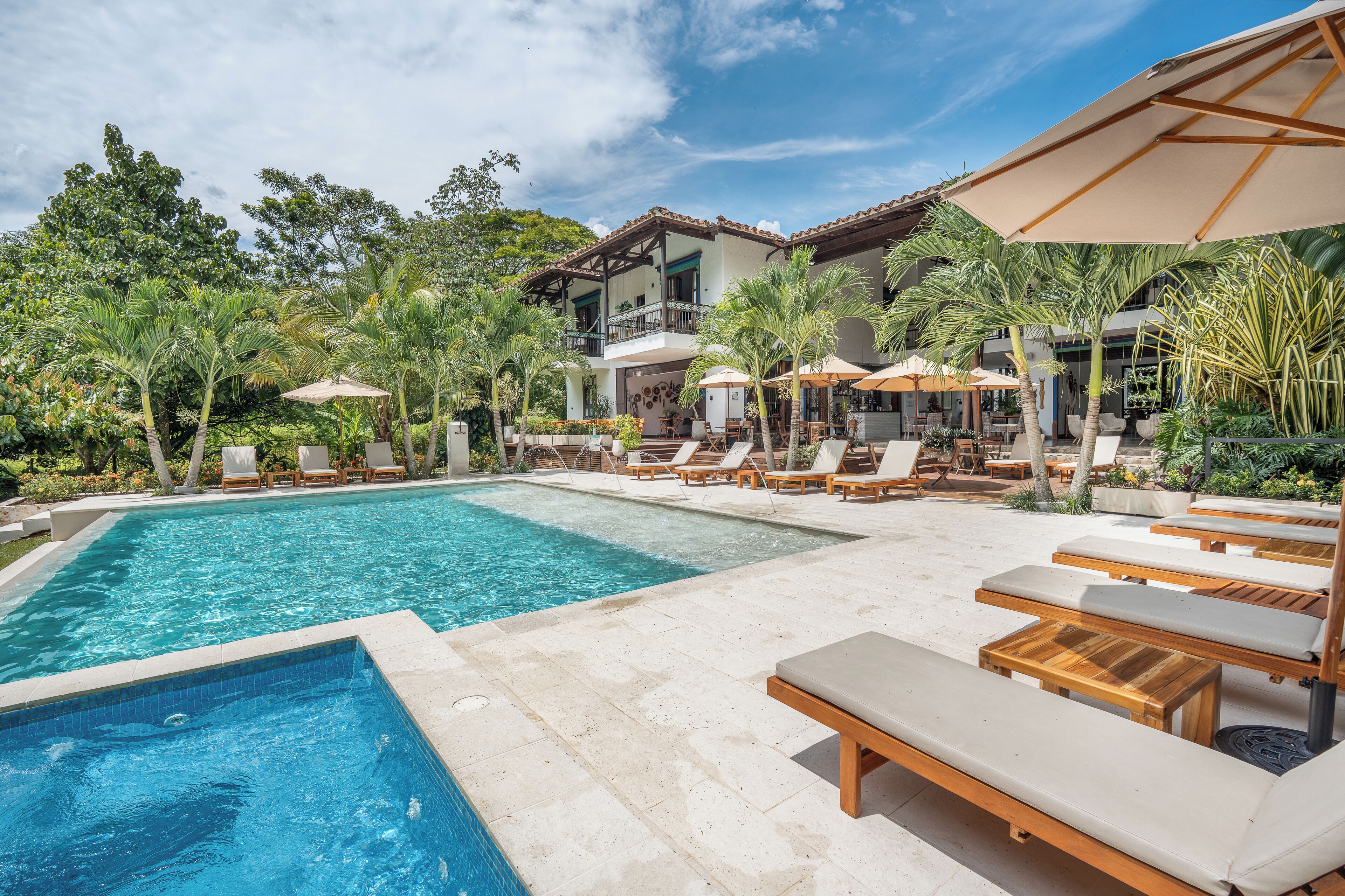 A pool area with lounge chairs and umbrellas, surrounded by palm trees and a building with balconies.