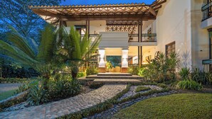 Una casa de dos pisos con un patio, un camino de piedra y plantas tropicales.