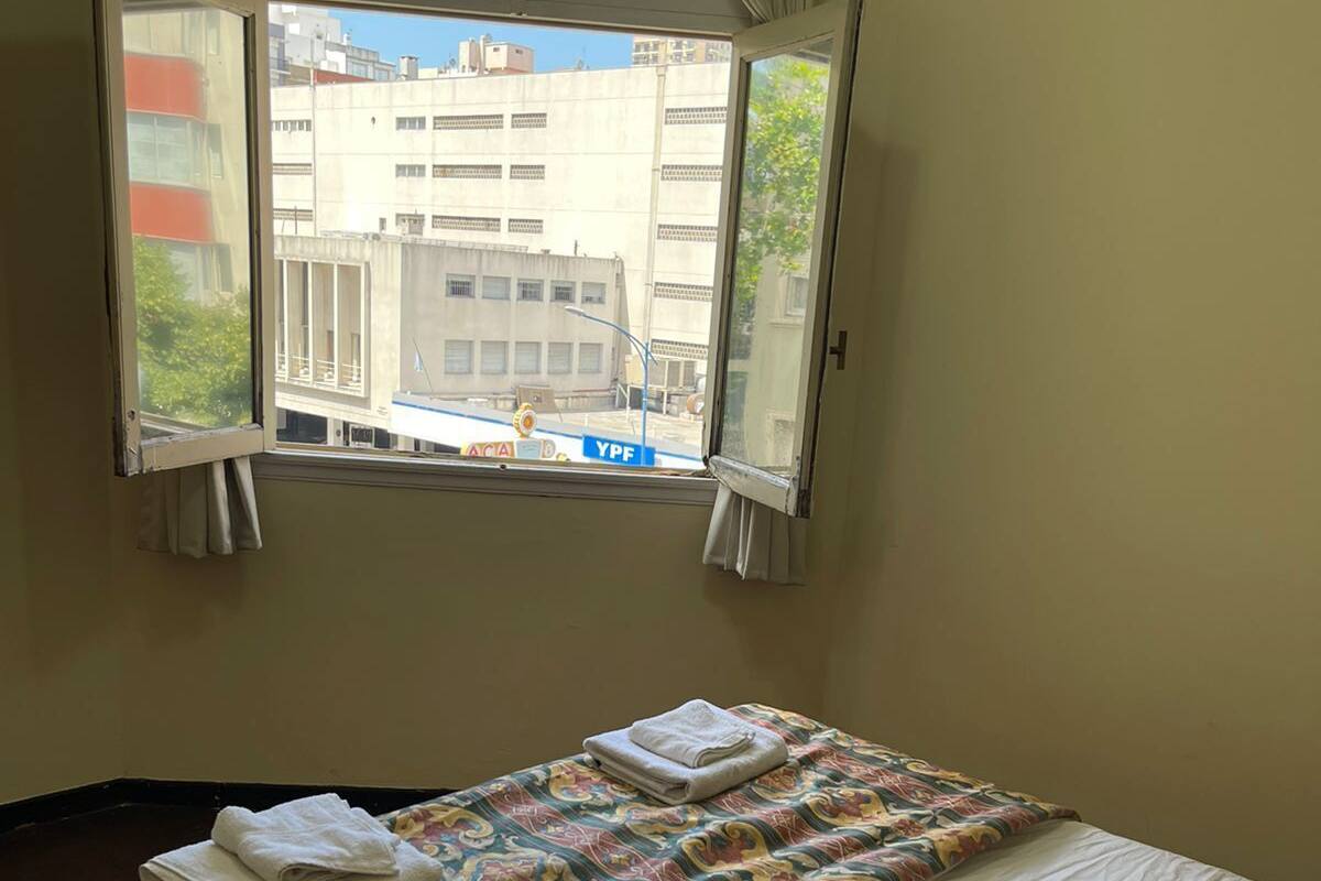 A single bed with a patterned bedspread and white sheets, a window with curtains, and a view of buildings outside.