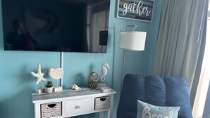 A living room with a teal wall, a flat-screen TV, a white console table with decorative items, a blue sofa with a 'Beach Life' pillow, and a 'gather' sign on the wall. - HS-0507 Oceanfront 1-BR Condo in Myrtle Beach, 652 sq ft of coastal comfort for up to 6 guests. (Myrtle Beach)