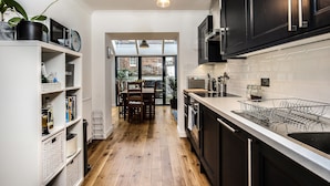 Cozinha moderna com armários pretos, bancada branca e piso de madeira. É possível ver uma área de jantar com mesa e cadeiras através de uma porta aberta.