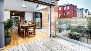 Uma casa moderna com deck de madeira, área de refeições com mesa e cadeiras, e uma grande porta de vidro que dá para um jardim.