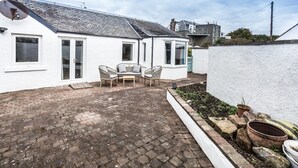 A white house with a tiled patio, two windows, and a small garden area with potted plants. - A delightful, homely cottage.  A great base for all of East Lothian. Sleeps 4. (Gullane)