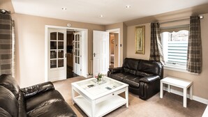 A living room with a leather sofa, a white coffee table, and a small white side table. There is a door leading to a kitchen and a window with curtains. - A delightful, homely cottage.  A great base for all of East Lothian. Sleeps 4. (Gullane)