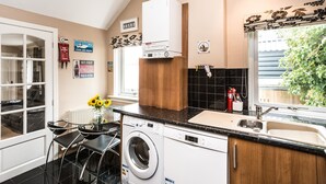 A compact kitchen with a built-in washer and dryer, a dining area with a table and chairs, a window with a view of greenery, and a decorative wall with signs. - A delightful, homely cottage.  A great base for all of East Lothian. Sleeps 4. (Gullane)