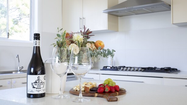 A kitchen counter with a bottle of wine, two wine glasses, a fruit platter, and a floral arrangement. - Cheerful Cottage Perfect for groups - Pool Table, Fire pit, Wifi & AC (Cessnock)
