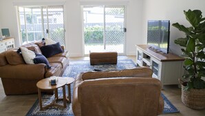 A living room with a brown leather sofa, a wooden coffee table, and a TV stand with a flat-screen TV. There is a large window with a view of a fence and greenery outside. - Cheerful Cottage Perfect for groups - Pool Table, Fire pit, Wifi & AC (Cessnock)