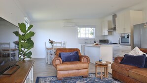 A modern kitchen with white cabinets, a stainless steel refrigerator, and a microwave. There is a dining area with a wooden table and chairs. - Cheerful Cottage Perfect for groups - Pool Table, Fire pit, Wifi & AC (Cessnock)