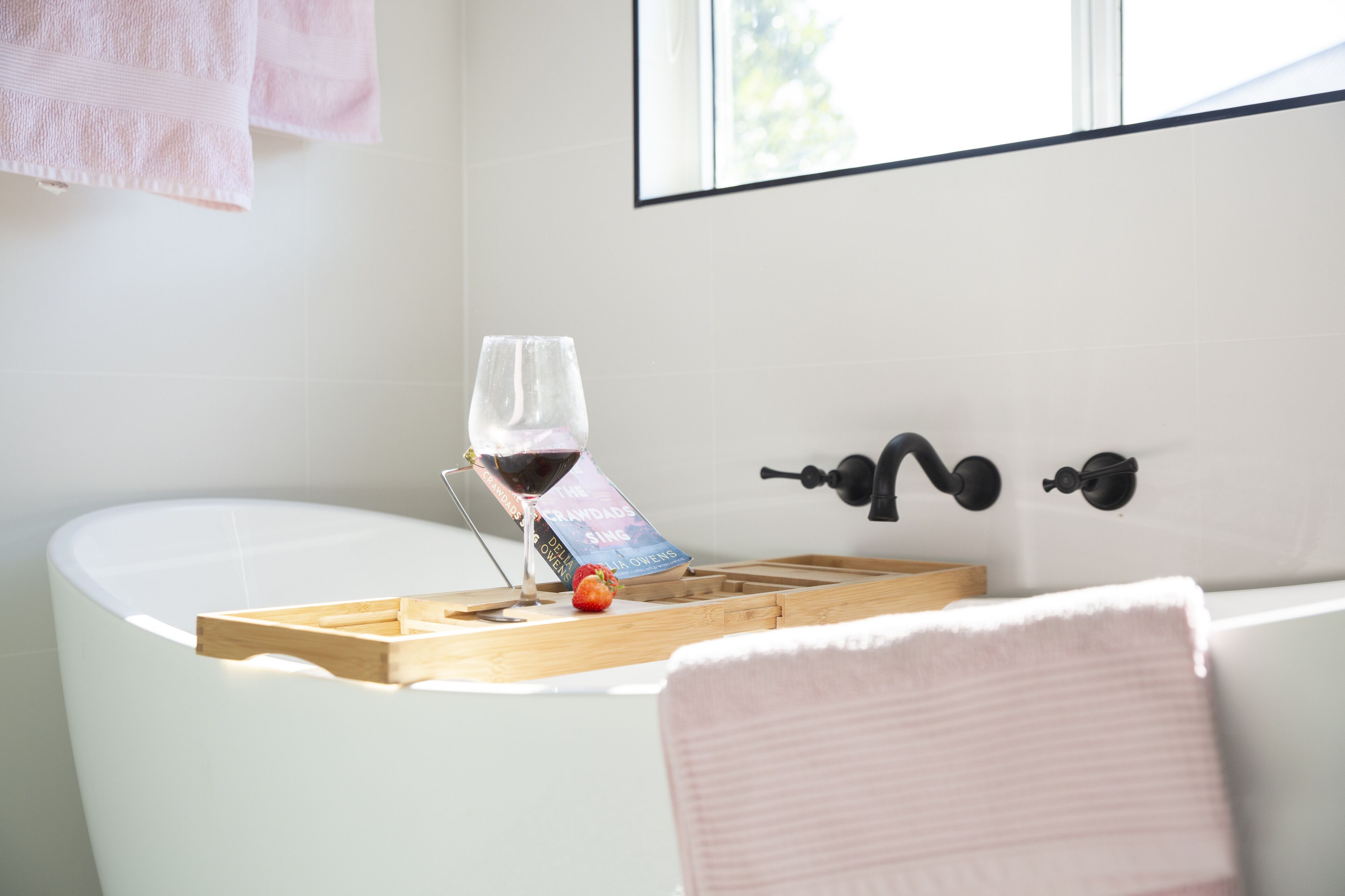A bathroom with a bathtub, a towel rack, a window, and a wine glass on a tray.