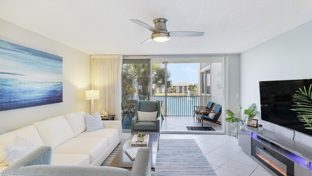 A modern living room with a white sofa, a grey armchair, a coffee table, and a flat-screen TV. There is a ceiling fan, a large painting, and a view of the outdoors through a sliding glass door. - Endless Summer,Jupiter Walk to Beach & Restaurants! (Jupiter)