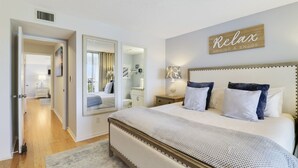 A bedroom with a large bed, a mirror, a bedside table with a lamp, and a 'Relax' sign on the wall. - Endless Summer,Jupiter Walk to Beach & Restaurants! (Jupiter)