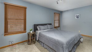 Ein Schlafzimmer mit einem Bett, zwei Fenstern mit Jalousien, einem Nachttisch mit Pflanze und einer Deckenleuchte.
