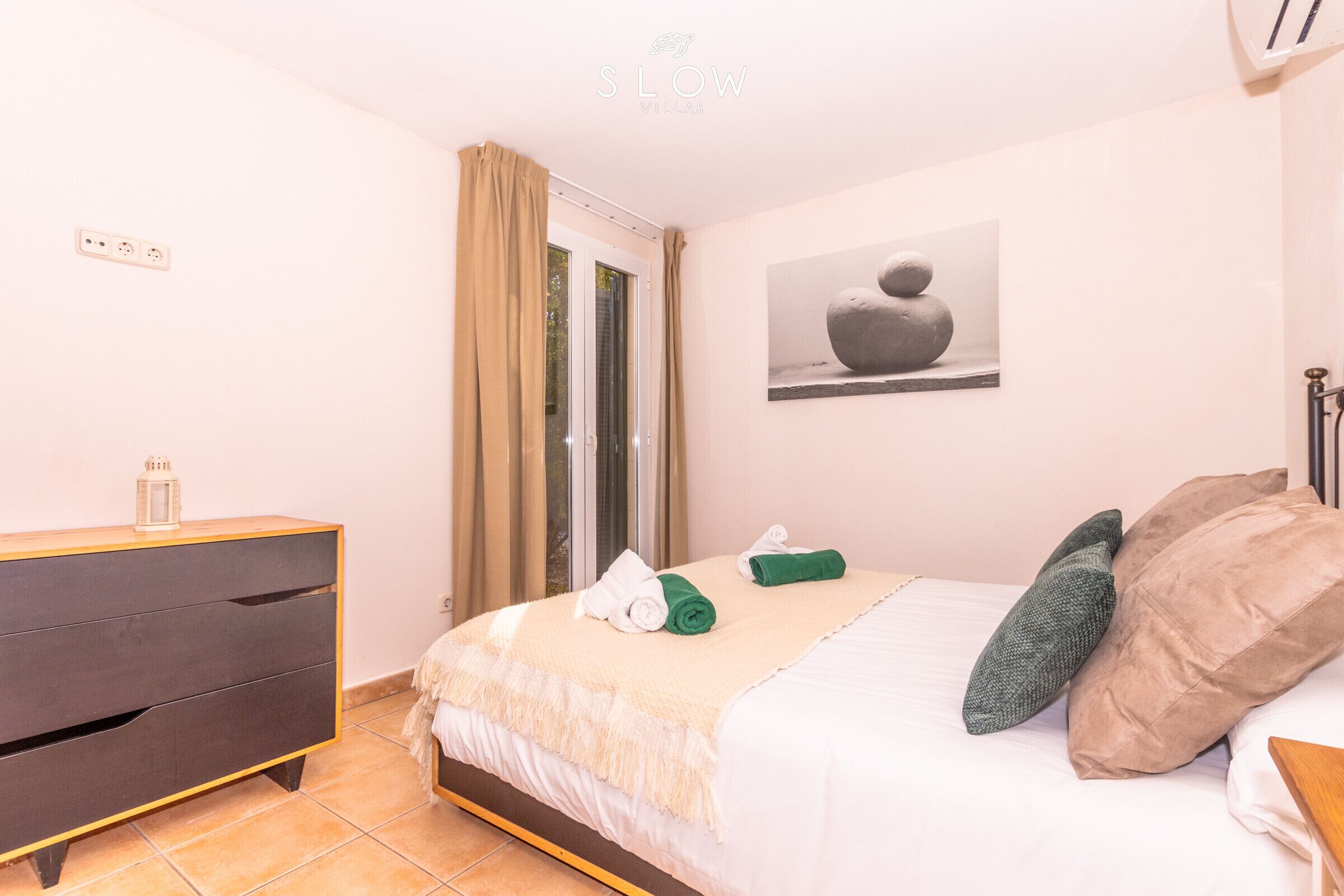 A bedroom with a bed, a dresser, a window with curtains, and a wall-mounted artwork.