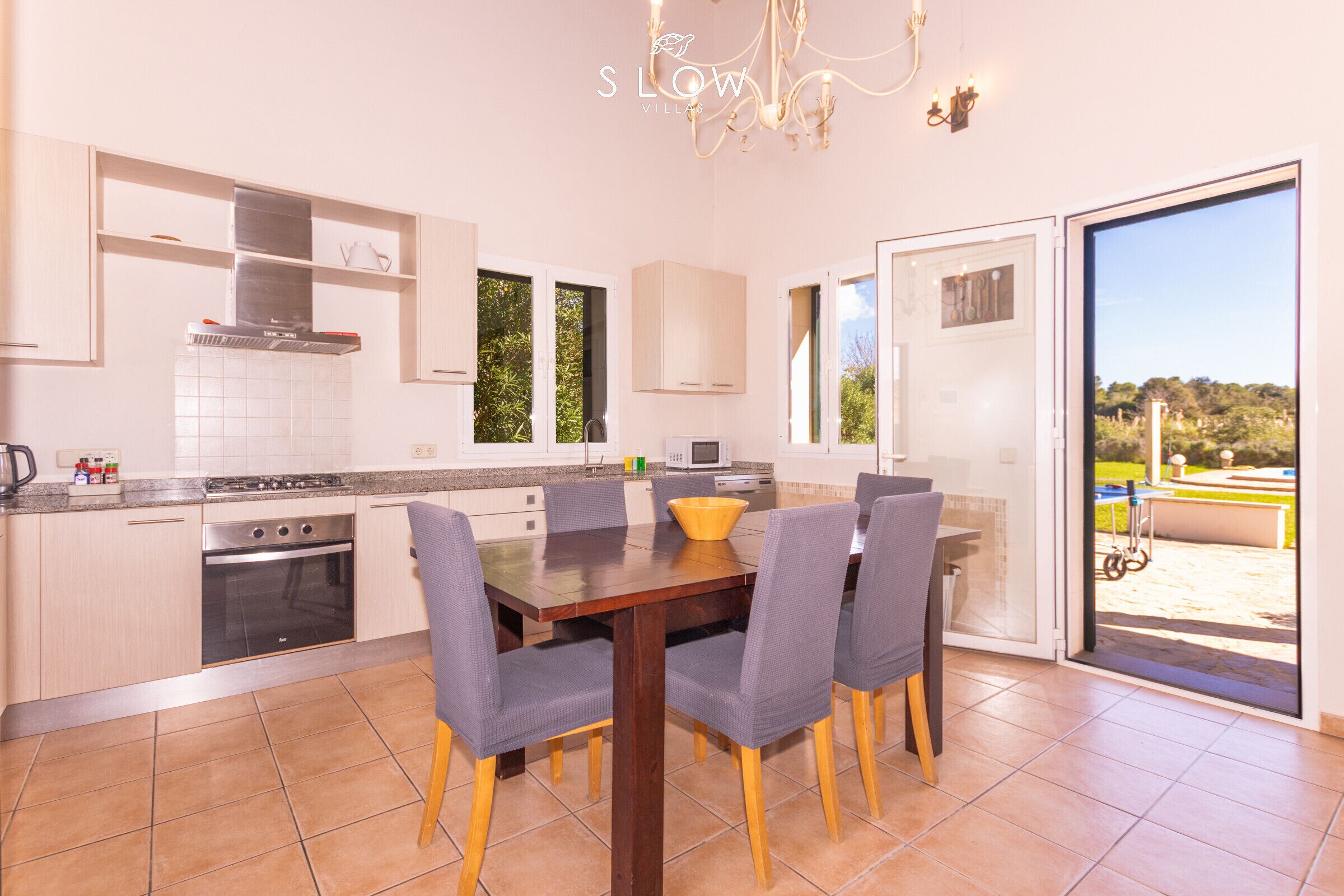 A modern kitchen with a dining area, featuring a large window, a chandelier, and a view of the outdoors.