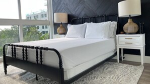 A modern bedroom with a black metal bed frame, white bedding, and a herringbone-patterned wall. There are two bedside tables with lamps and a view of the city through large windows. - Your Bay View Escape in Coconut Grove, Pool & Gym (Miami)