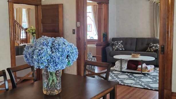 A dining area with a wooden table, chairs, and a vase of blue flowers. There is a sofa and a small round table with a basket in the background.