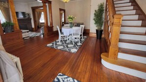 A spacious living room with hardwood floors, a dining area with a white table and chairs, a staircase with wooden railings, and a chandelier.