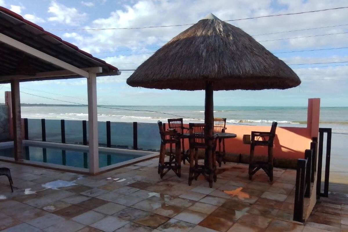A beachside patio with a thatched umbrella, a pool, and wooden chairs.
