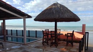 A beachside patio with a thatched umbrella, a pool, and wooden chairs.