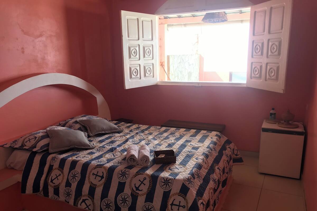 A bedroom with a bed, pillows, a bedside table, a nightstand, and a window with open shutters.