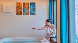 Une personne est assise près d’une fenêtre, dans une pièce qui contient un jacuzzi, trois tableaux accrochés au mur et une serviette posée sur un banc.