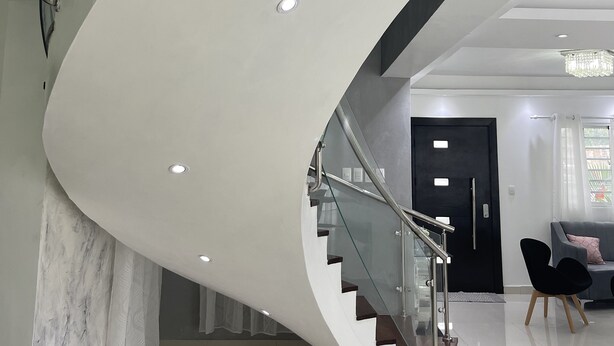 A modern spiral staircase with glass railings and a sleek design, leading to a well-lit living area with a sofa and chairs. - 5 minutes from la caleta beach 10 minutes from casa de campo la romana (romana del este)