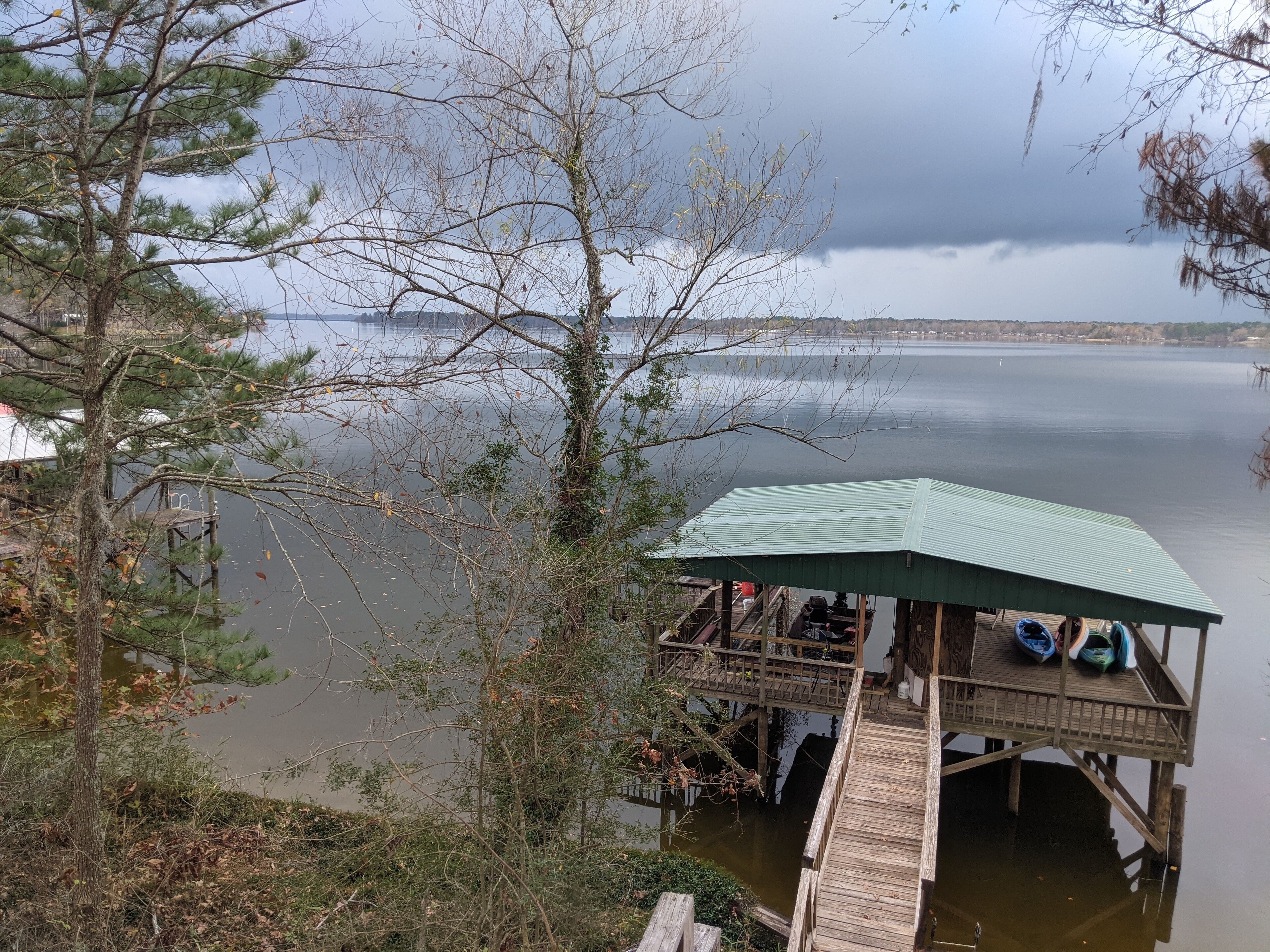 A wooden dock with a covered seating area overlooking a calm lake, surrounded by trees and a distant shoreline.