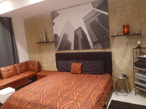 A bedroom with a bed, a brown leather sofa, a patterned orange bedspread, a cityscape artwork, a small table with a lamp, and a shelf with decorative items. - Mr. Whips Family Fun House 3,000 Sq Ft Game unit with arcade room and rear yard (Philadelphia)