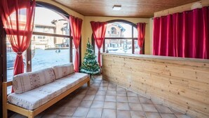 Une pièce chaleureuse avec un banc en bois, des rideaux rouges, un petit sapin de Noël et une vue sur les bâtiments à l’extérieur.