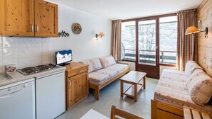 Une cuisine compacte avec des meubles en bois, un lave-vaisselle blanc, une cuisinière et une petite table. On y trouve également un canapé et une fenêtre donnant sur les arbres.