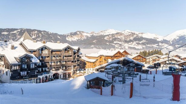 Une station de montagne enneigée, avec de nombreux bâtiments en bois, des remontées mécaniques et une vue dégagée sur les montagnes en arrière-plan.