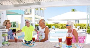 A group of people at an outdoor pool area with a table, drinks, and a blender.