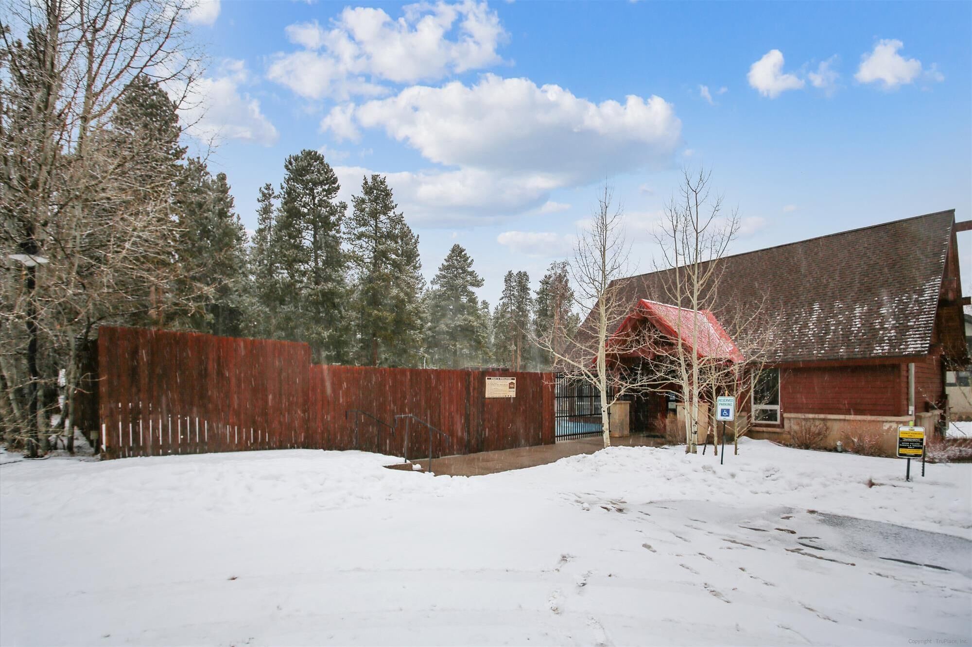 A snow-covered parking area with a wooden fence, a building with a red roof, and a sign indicating handicapped parking.
