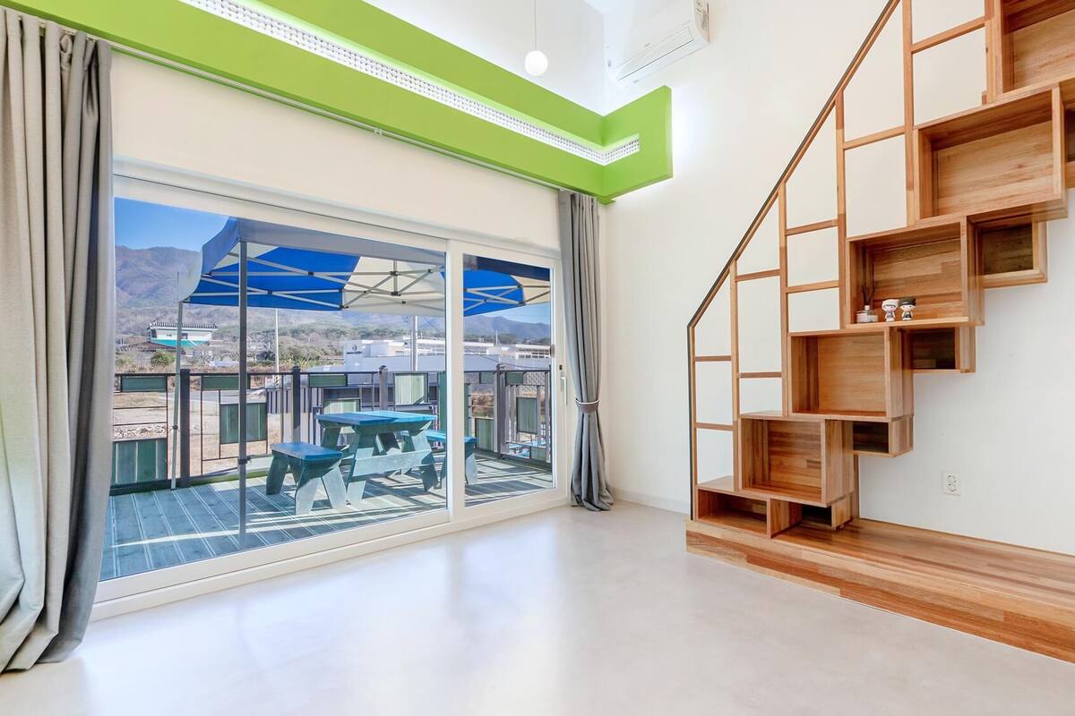 Ein modernes Interieur mit einer großen Schiebeglasdoor, die zu einer Terrassenanlage führt, einer Holztreppe mit integrierten Regalen und einer grünen Akzentwand.