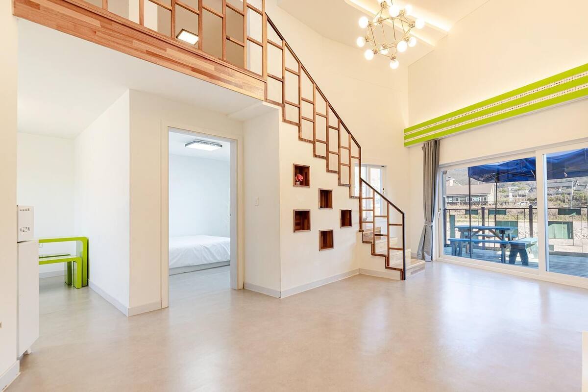 Ein geräumiger Raum mit einer Holztreppe, einem großen Fenster mit Blick auf eine Sitzgelegenheit im Freien und einer hellen Deckenleuchte.