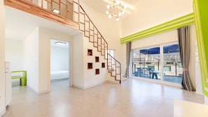 Une pièce spacieuse avec un escalier en bois, une grande fenêtre donnant sur un espace extérieur aménagé, et un luminaire au plafond qui illumine l’ensemble.