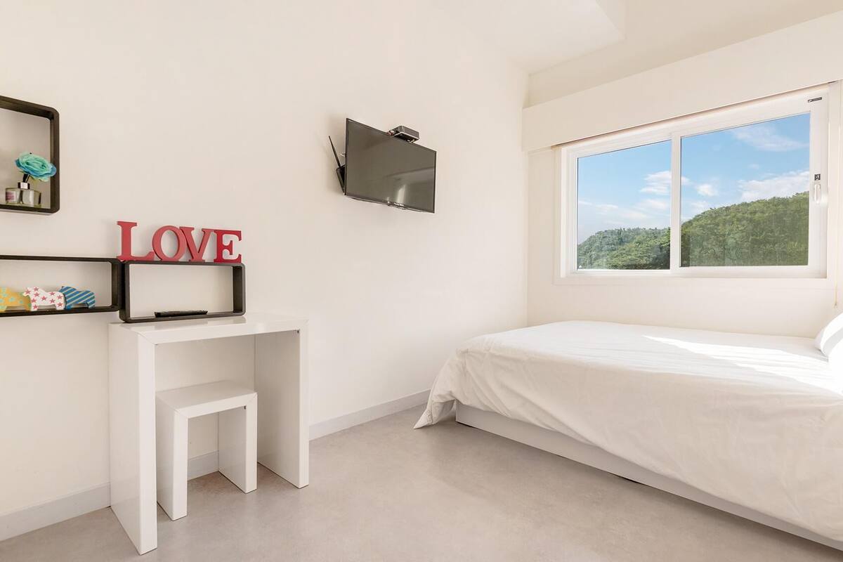 Ein Schlafzimmer mit Bett, Schreibtisch, Fernseher, Fenster mit Blick auf Grünflächen und einem 'LOVE'-Schild an der Wand.