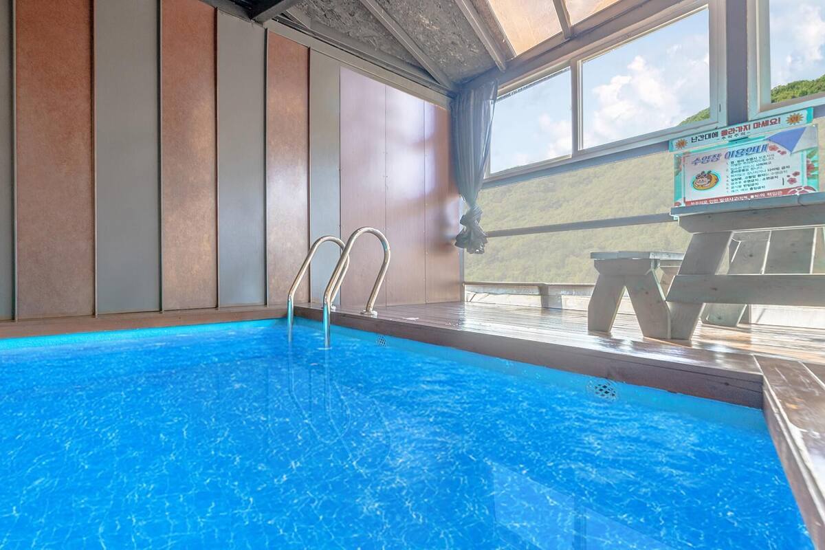 Ein Hallenbad mit Metallleitern, einer Holzterrasse und Blick ins Freie.