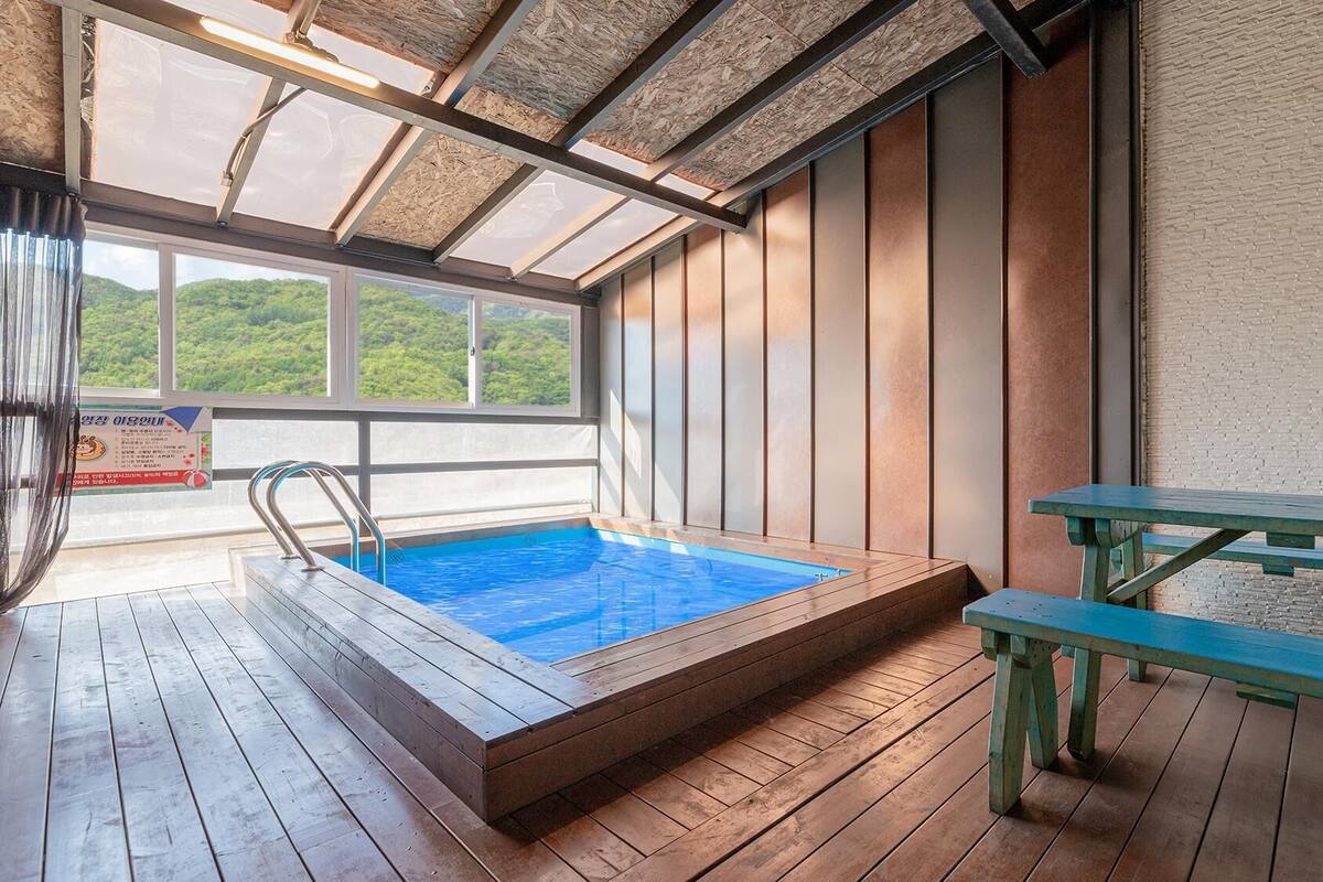 Eine Holzterrasse mit Whirlpool und Picknicktisch, große Fenster mit Blick auf Grünflächen und ein Schild in koreanischer Schrift.