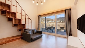 Ein geräumiges Wohnzimmer mit grauem Sofa, einer Holztreppe und einer großen Schiebeglastür, die zu einer Terrasse mit Blick auf die Hügel führt.