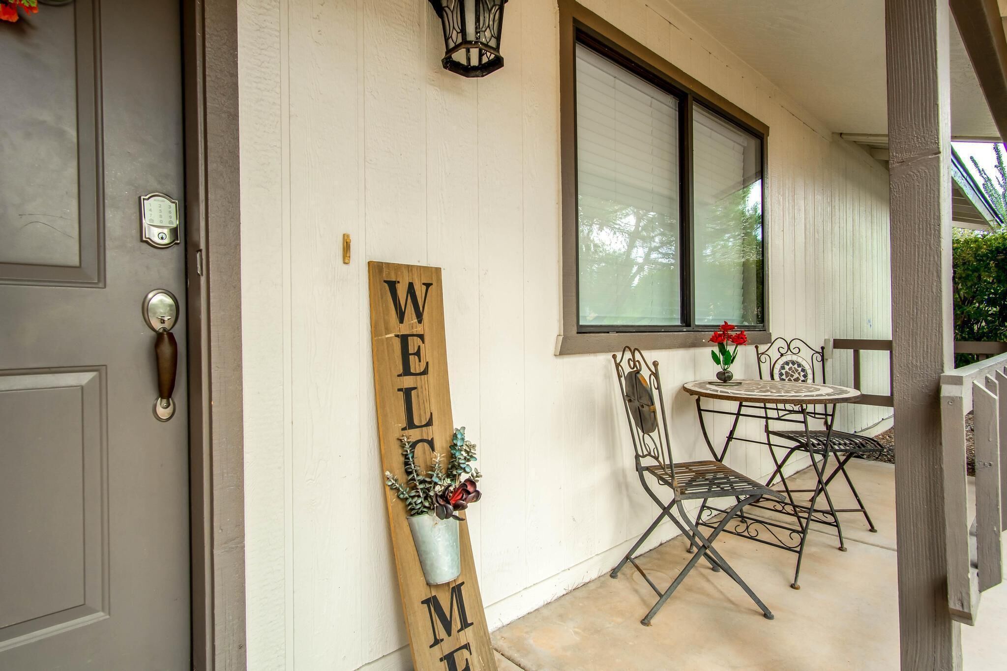 A wooden welcome sign with a potted plant, a metal folding chair, and a table with a vase of flowers on a porch.