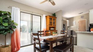 Un comedor con una mesa de madera, sillas y un ventilador de techo. Hay una puerta corrediza de vidrio que da a un balcón y una zona de cocina con armarios y electrodomésticos.