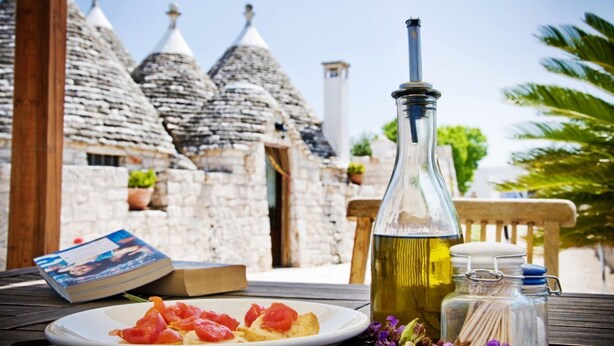 Une table avec une assiette de nourriture, une bouteille d’huile d’olive et un livre, le tout placé devant un bâtiment en pierre doté de toits coniques.