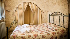 Une chambre avec un mur en pierre, un lit avec une literie à motifs floraux, une tête de lit en métal et des rideaux.
