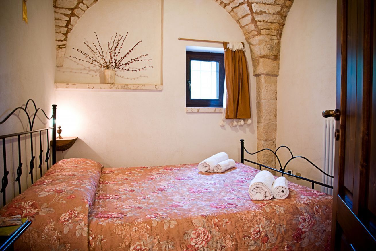 A bedroom with a bed, a window with curtains, a wall-mounted artwork, and a stone archway.